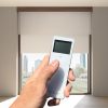 Hand clicking on a 15-channel, Fabtex handheld remote for roller shade. In the background are blackout & sheer shades.